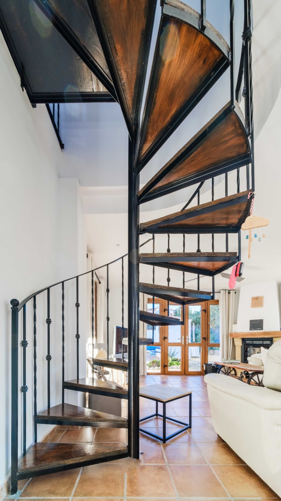 Spiral staircase with a black metal frame and dark wood treads, set within a living space featuring terracotta tiles and views of the garden.