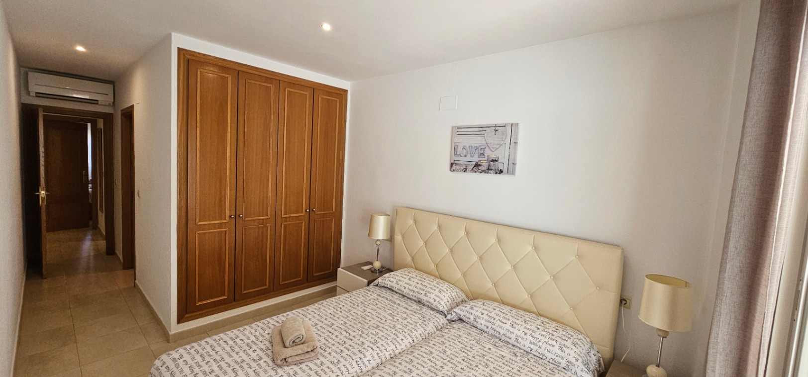 Bedroom featuring a four-door built-in wooden wardrobe, a cream tufted headboard, and recessed lighting with an air conditioning unit above the hallway door.