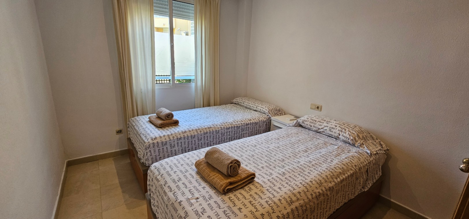 Bedroom featuring twin beds, light tile flooring, and a window providing natural light with white curtains.