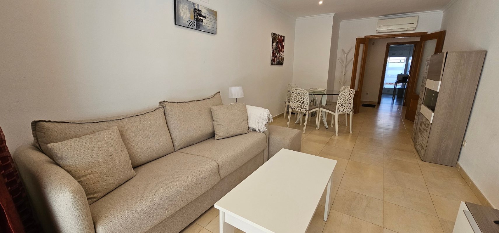 Bright living and dining area featuring a beige fabric sofa, white coffee table, and a glass dining set. The space includes tiled flooring and split-system air conditioning.
