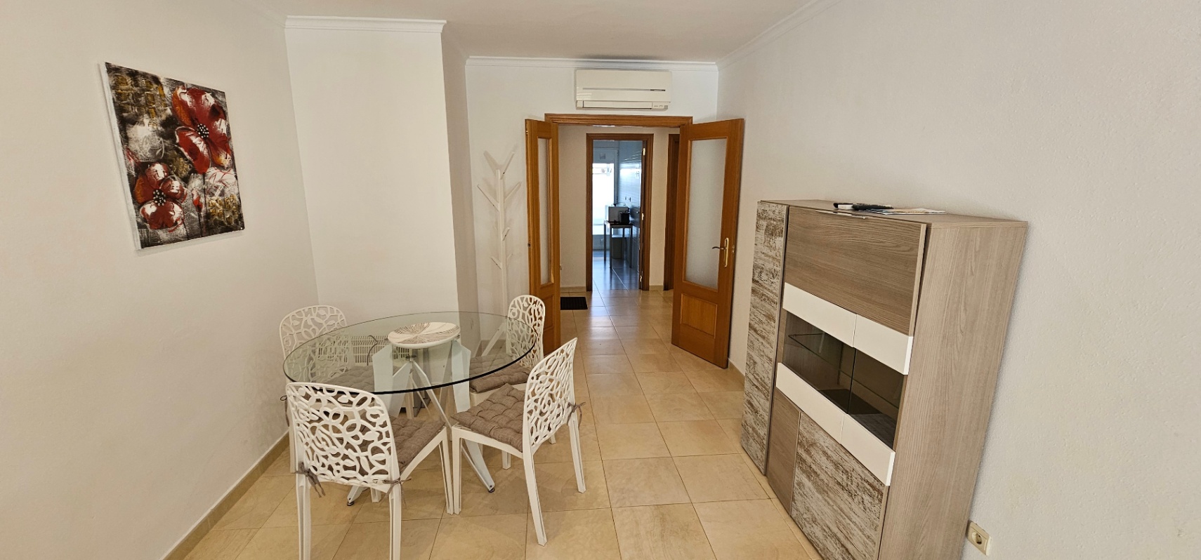 Dining area featuring a circular glass table with patterned white chairs and a modern sideboard, leading through wooden double doors to the hallway.