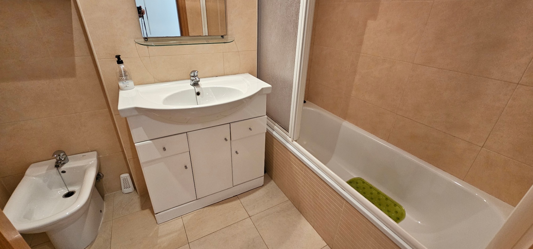 Full bathroom featuring beige ceramic tiling, a white vanity unit with storage, a bidet, and a bathtub with a glass partition.