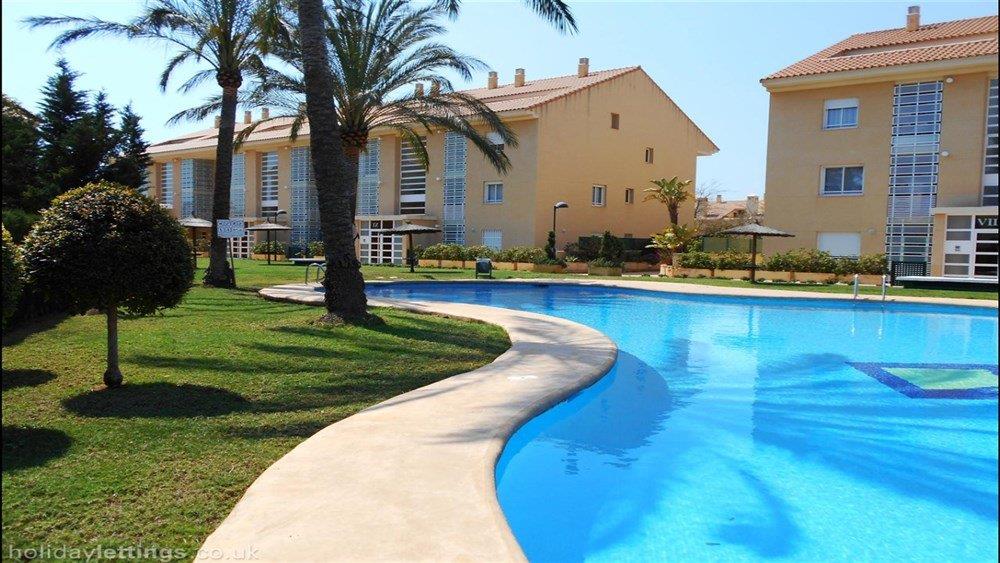 Large free-form community swimming pool featuring blue tiling, surrounded by manicured lawns, mature palm trees, and Mediterranean-style architecture.