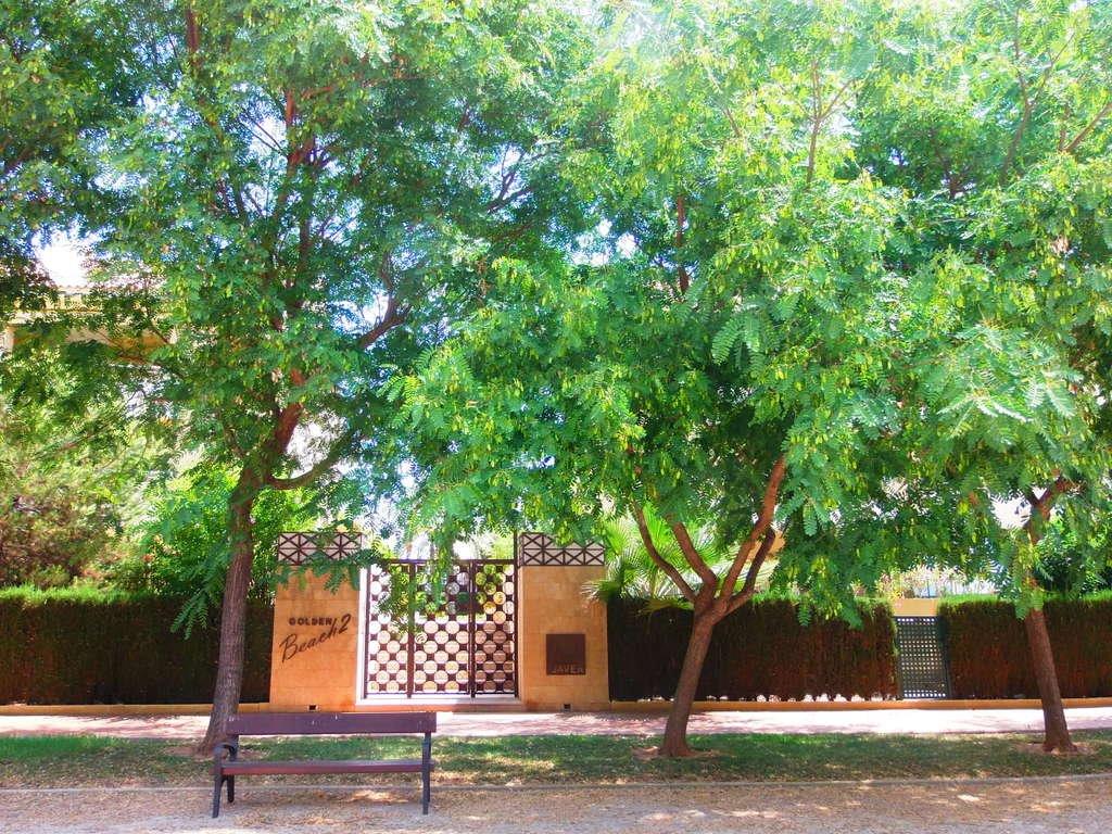 Main entrance of the Golden Beach 2 complex, featuring decorative iron gates, stone pillars, and mature trees providing shade over a wooden bench.