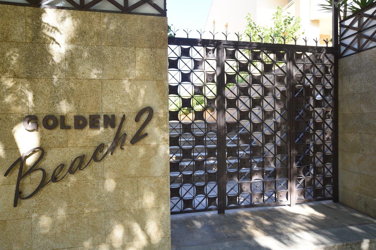 Main entrance to the Golden Beach 2 complex, featuring a textured stone wall with metal signage and a decorative geometric iron gate.