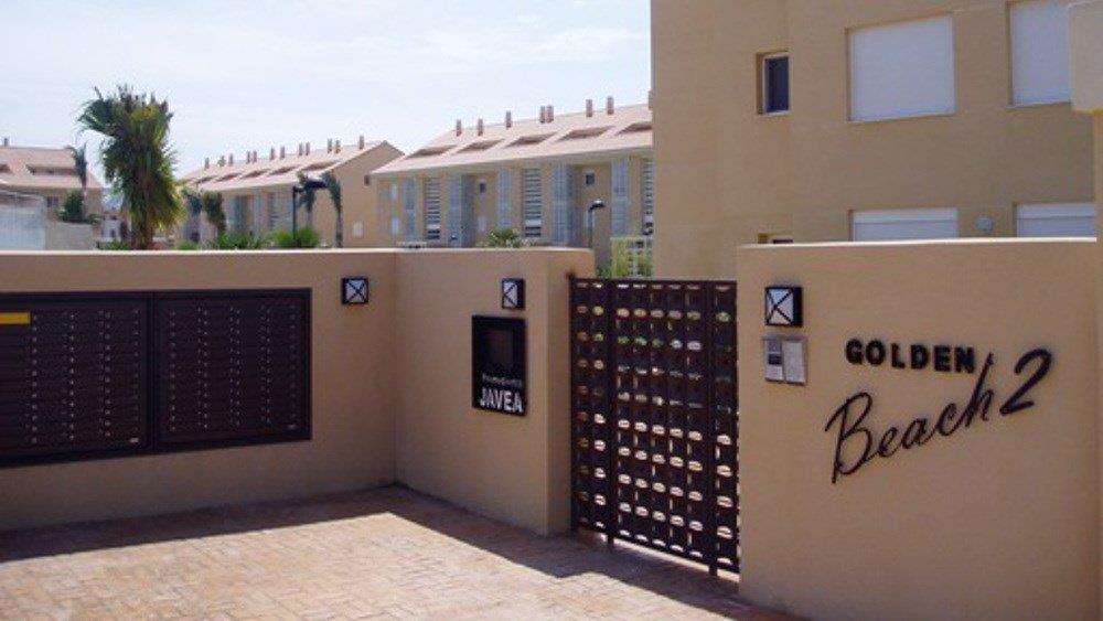 Main entrance to the Golden Beach 2 complex, featuring sand-colored walls, integrated mailboxes, and a dark metal gate under bright natural light.