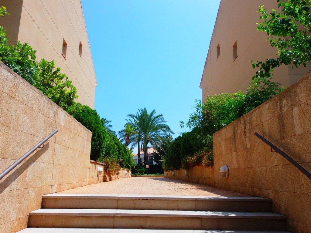 Outdoor entrance featuring stone steps and metal handrails, flanked by stone-clad walls and lush Mediterranean landscaping under a clear sky.