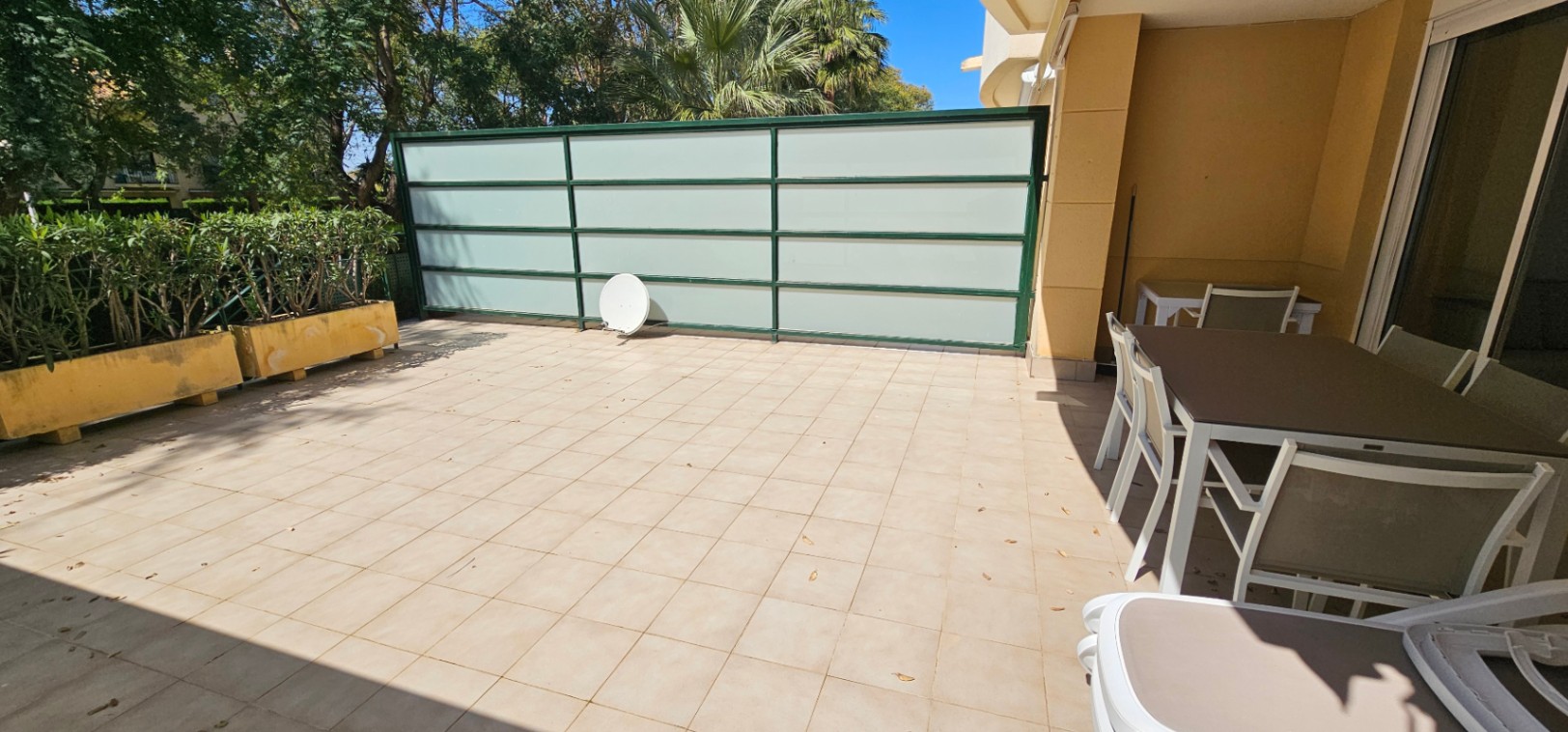 Private south-facing terrace featuring tiled flooring, an outdoor dining area, and privacy screens, bordered by planters and lush Mediterranean trees.