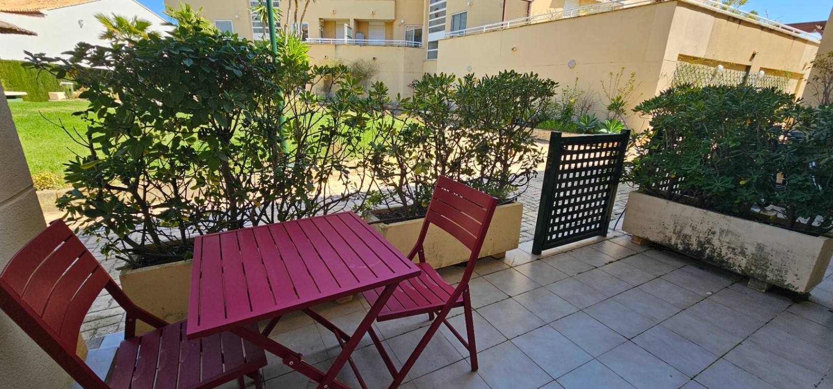 Private tiled terrace featuring burgundy wooden outdoor furniture and integrated planters, providing direct gated access to the communal gardens.
