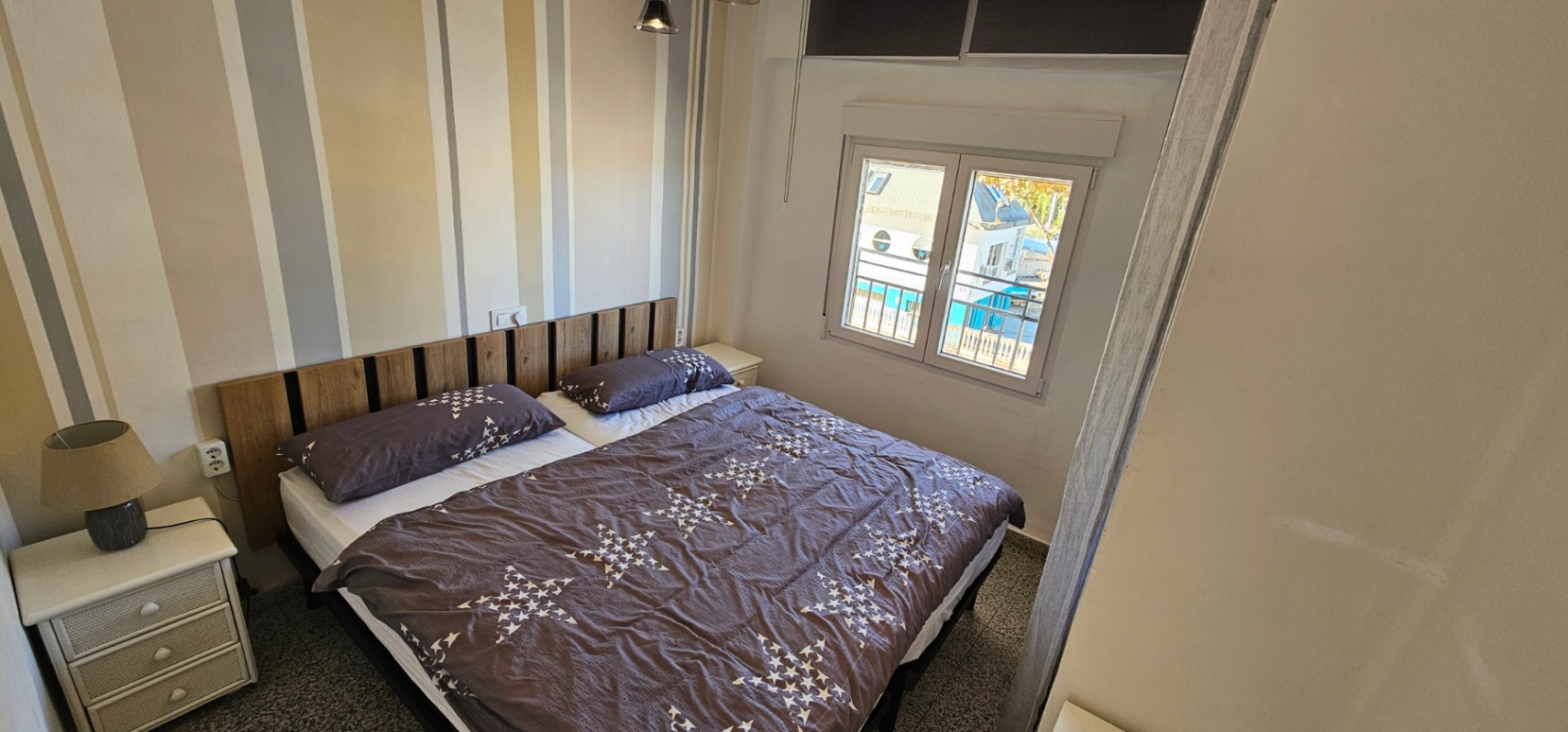 Bedroom featuring a double bed with a wooden slat headboard and neutral striped wallpaper. A white-framed window provides natural light and views of the surroundings.