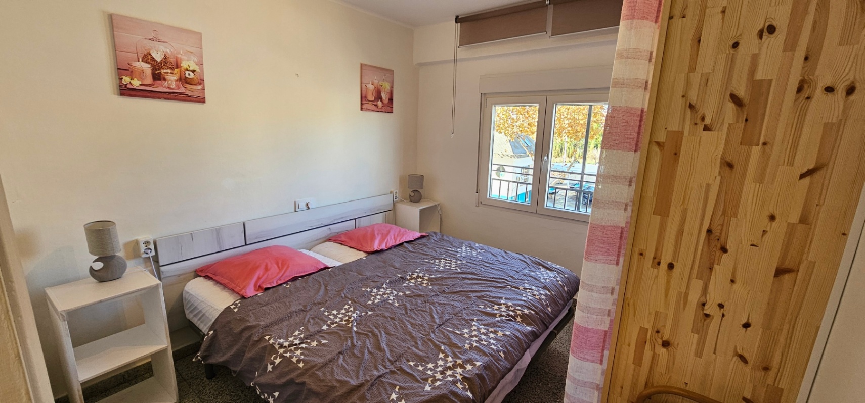 Bright bedroom featuring a double bed with a light wood headboard and white bedside shelving. A large window provides natural light and views of the neighborhood.