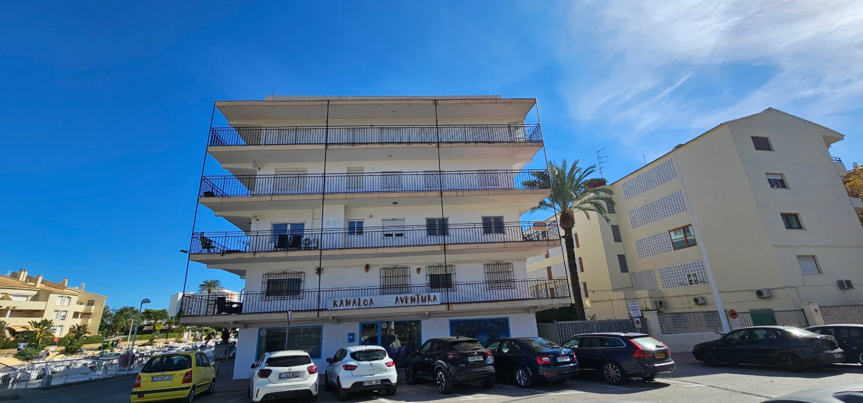 Exterior view of the Mediterranean-style building featuring wrap-around balconies with black railings and a commercial ground floor under a clear sky.