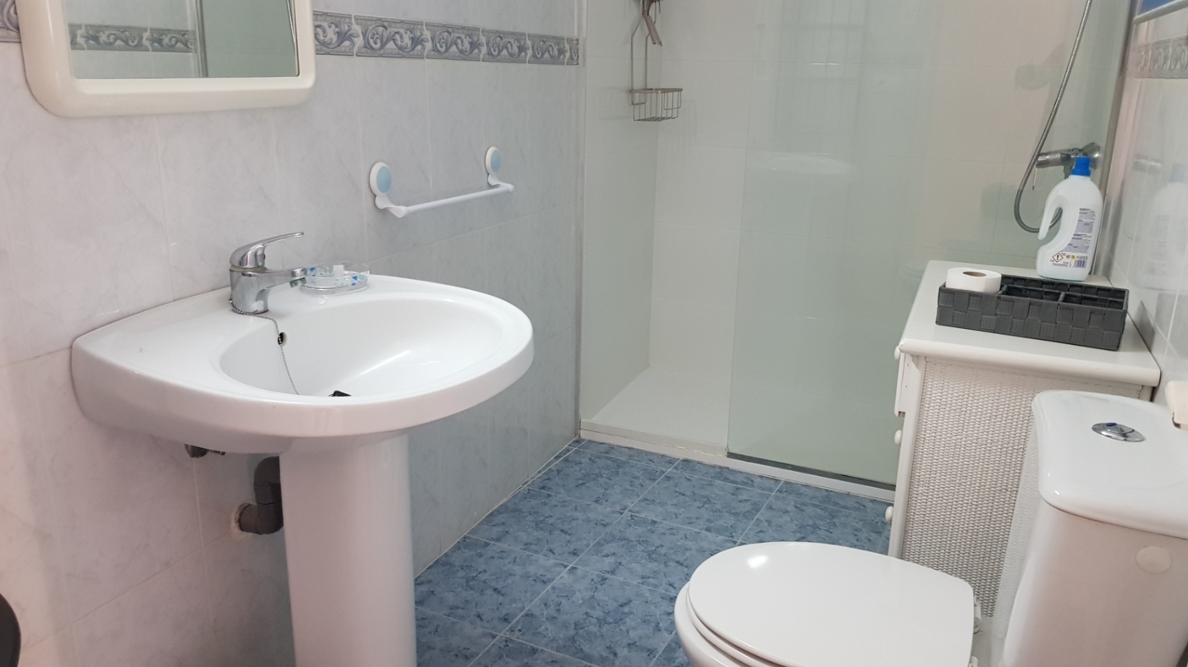 Functional bathroom featuring a pedestal sink, toilet, and walk-in shower with a glass screen. It includes blue floor tiles and a white wicker storage unit.