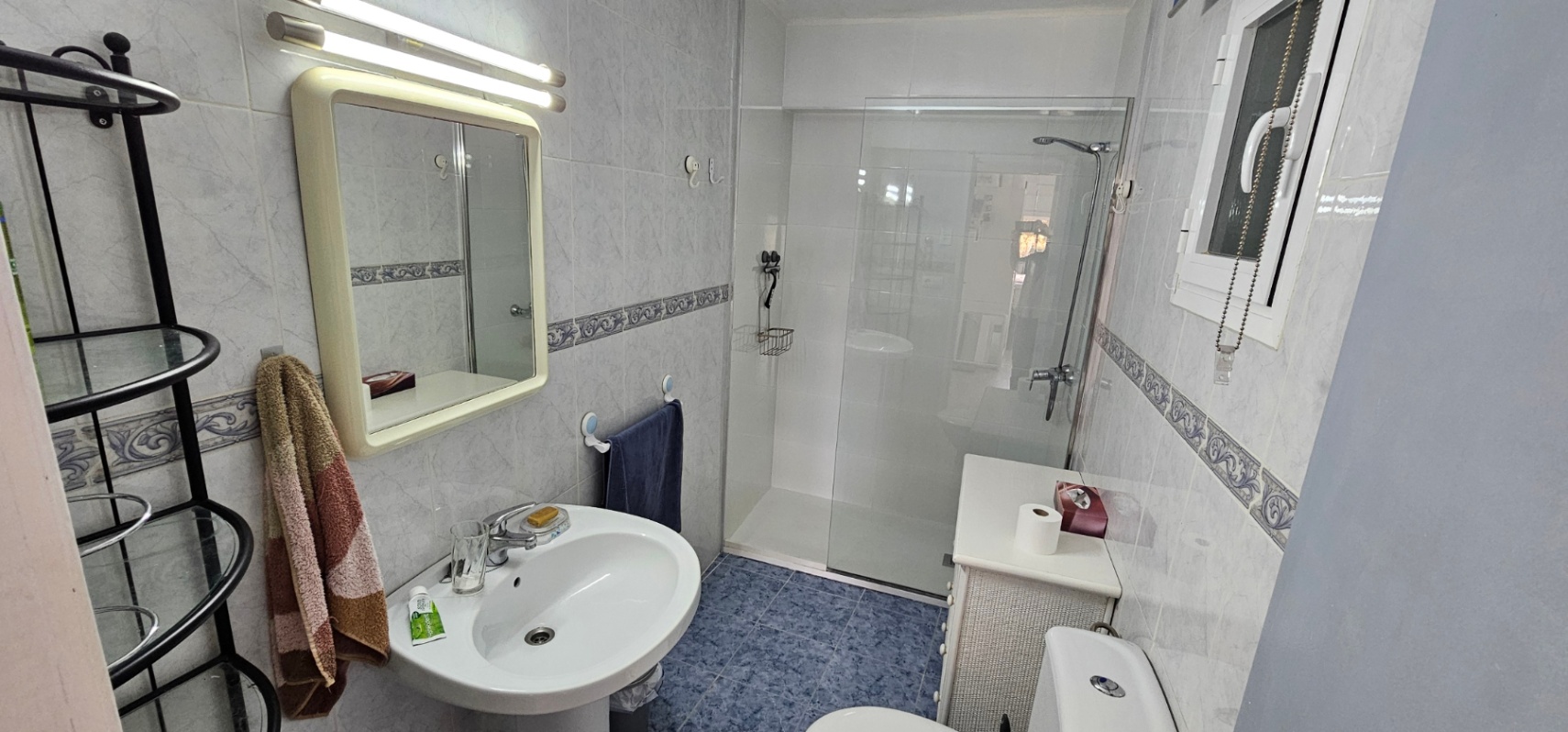 Functional bathroom featuring a walk-in shower with glass screen, pedestal sink, and blue floor tiles complemented by decorative wall borders.