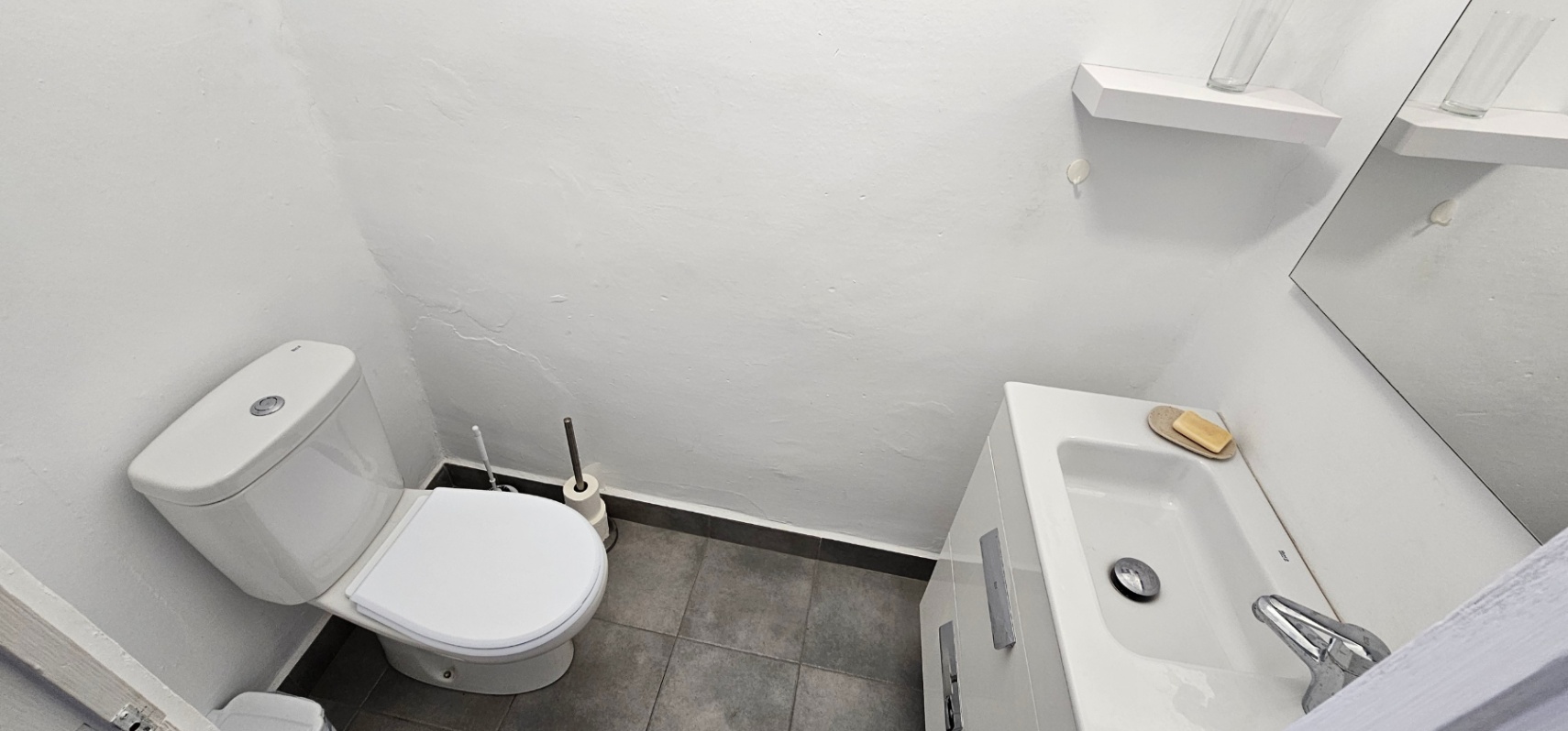 Functional bathroom featuring a white toilet, a vanity unit with an integrated sink, and grey floor tiles. The space includes a wall-mounted mirror and a small white shelf.