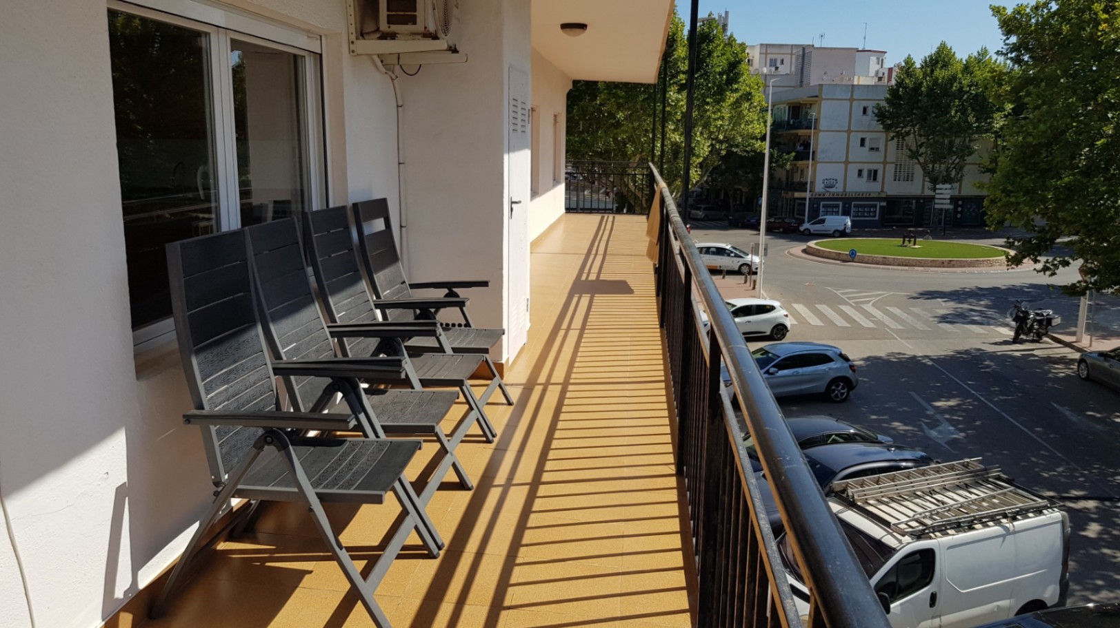 Long balcony featuring ochre floor tiles, grey folding chairs, and elevated views of the surrounding urban area and local greenery.
