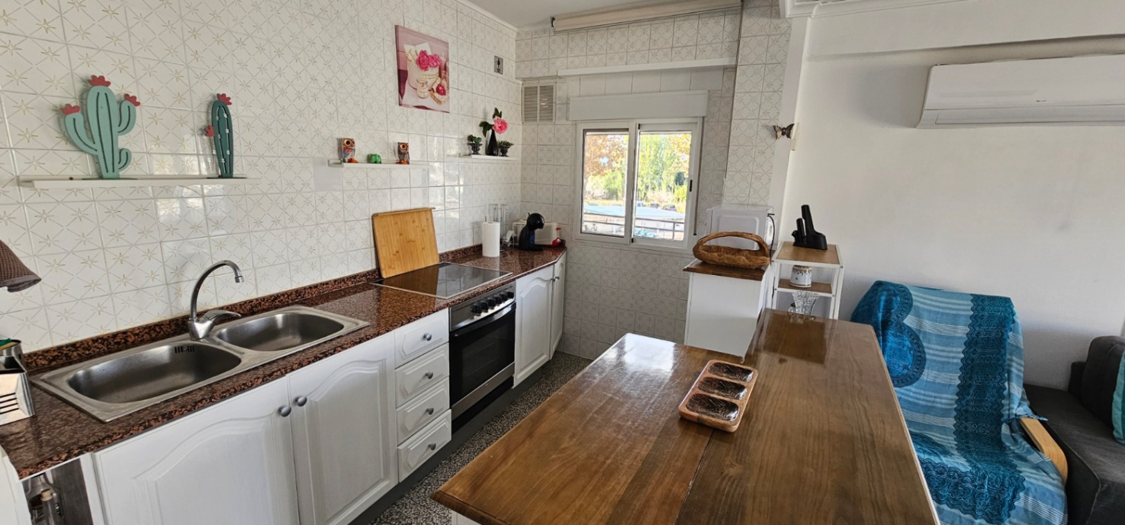 Open-plan kitchen featuring granite countertops, a double stainless steel sink, and white cabinetry. It includes a wooden dining table and wall-mounted air conditioning.