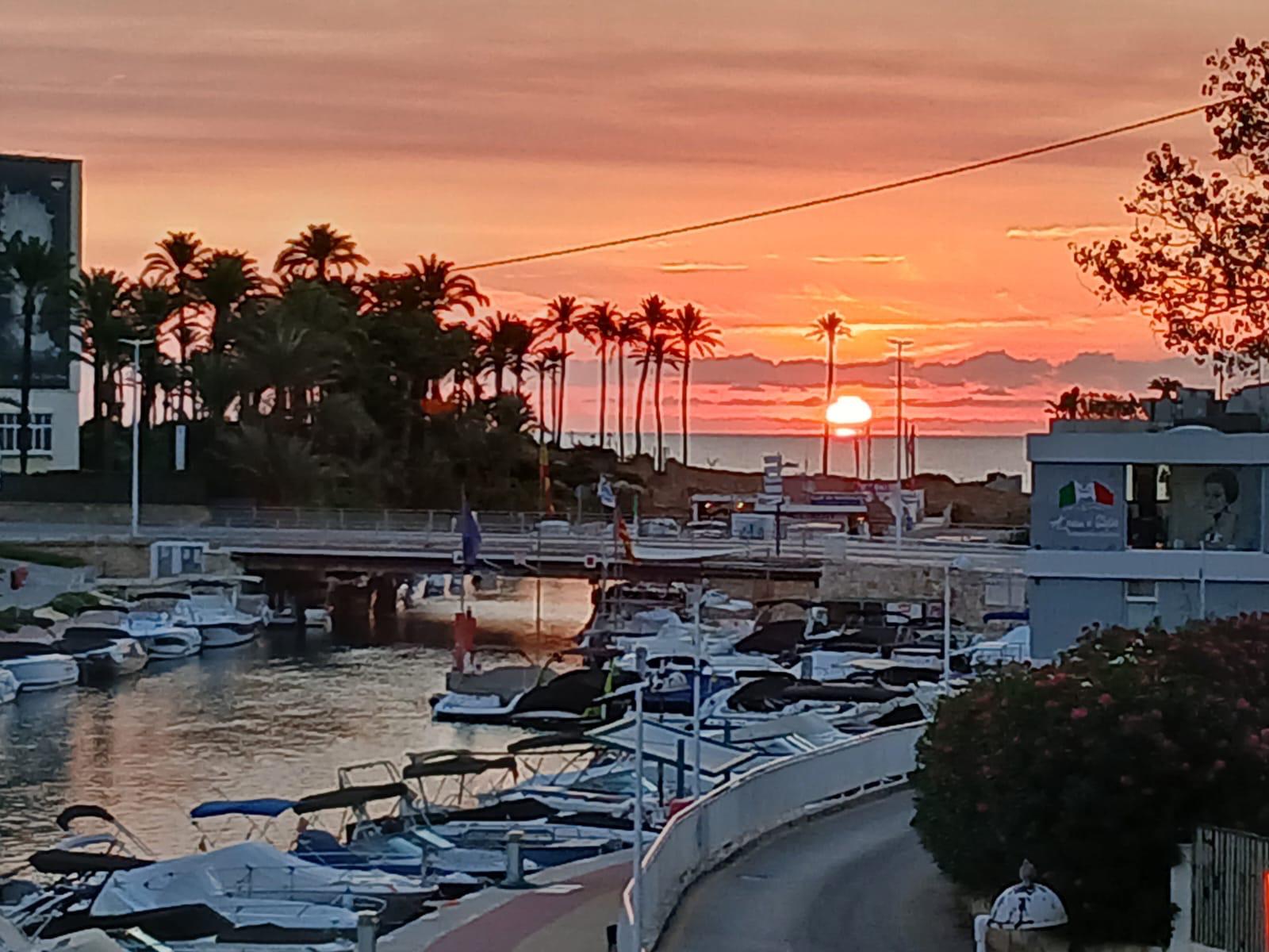 Panoramic sunset view from the terrace overlooking the Fontana canal, featuring moored boats, a bridge, and silhouetted palm trees against the Mediterranean horizon.