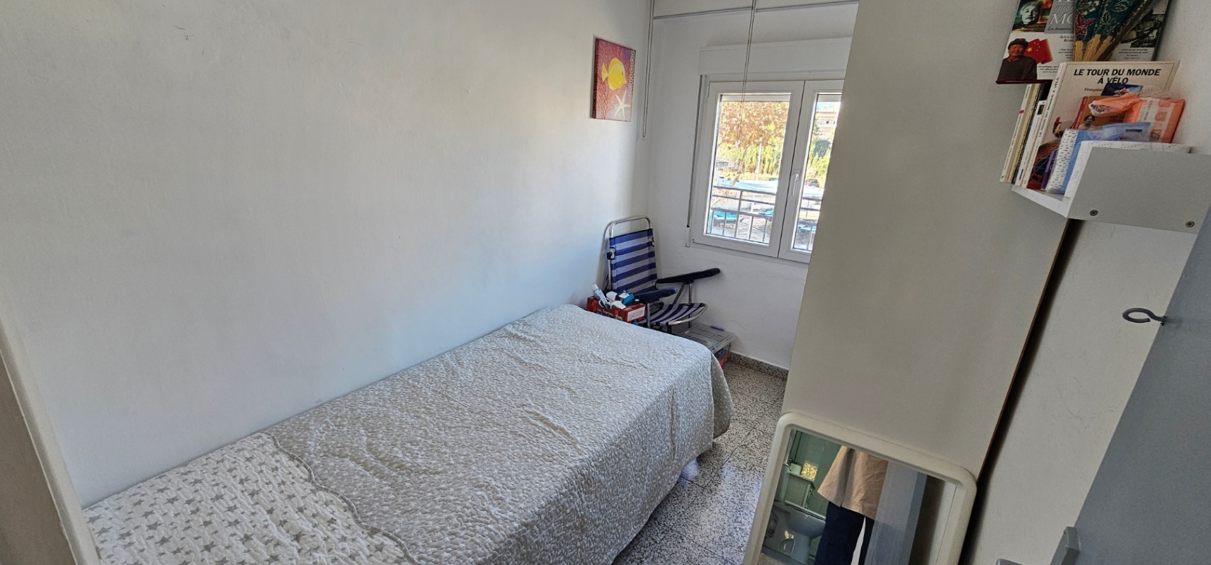 Single bedroom featuring a twin bed, original terrazzo flooring, and a window providing natural light. The space includes white walls and functional shelving.