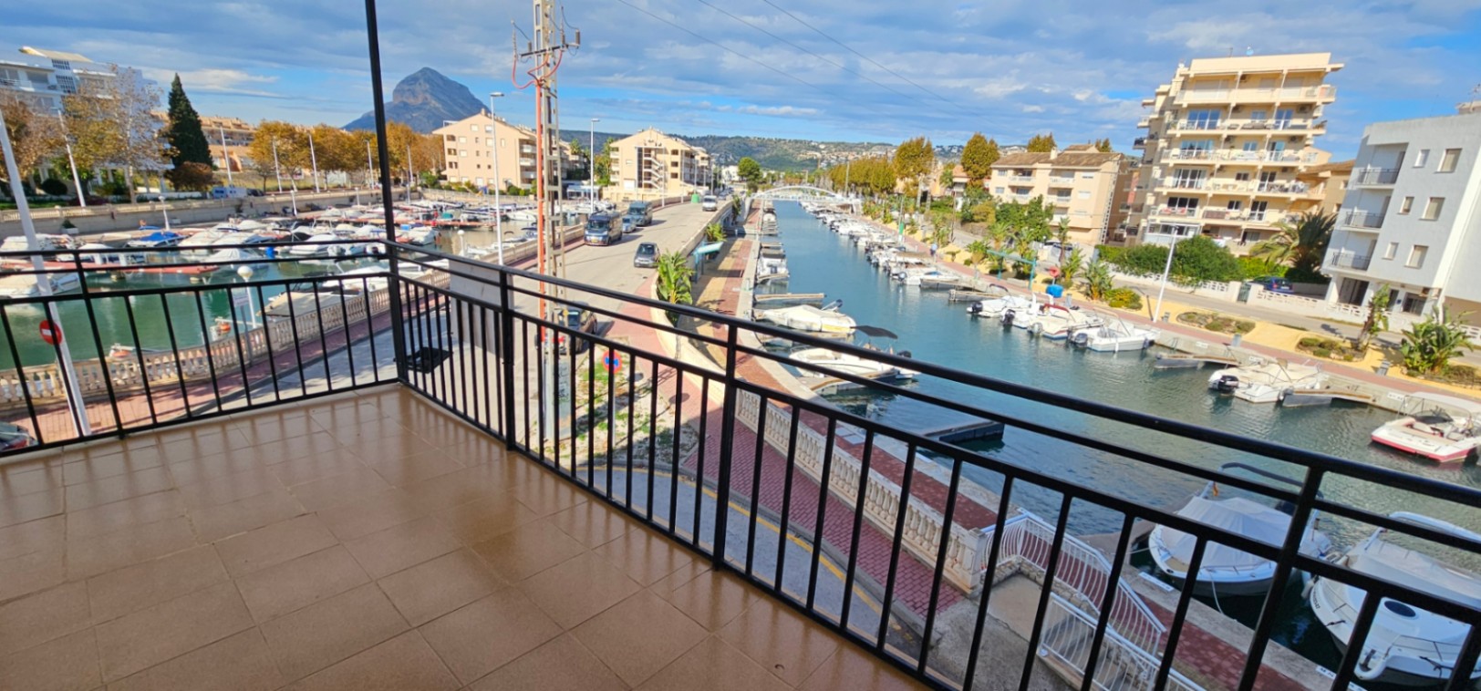 Tiled terrace with black metal railings overlooking the marina canal, featuring views of docked boats and the Montgó mountain in the distance.