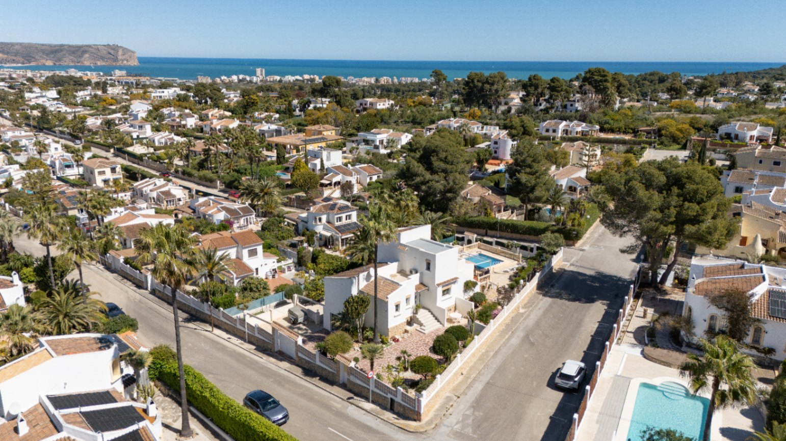 Aerial perspective of the white villa on a corner plot in Pinosol, Jávea. The image shows the private pool, landscaped garden, and panoramic views of the Mediterranean coastline.