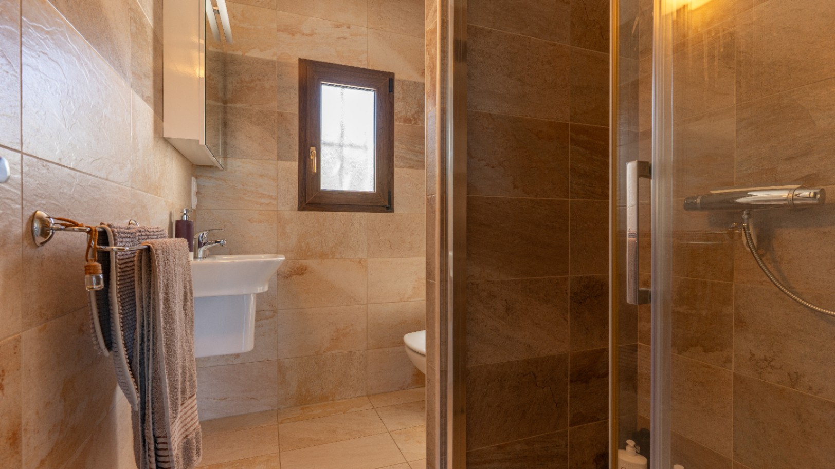 Bathroom featuring stone-effect ceramic tiling, a glass-enclosed walk-in shower, white pedestal sink, and a small wooden-framed window.