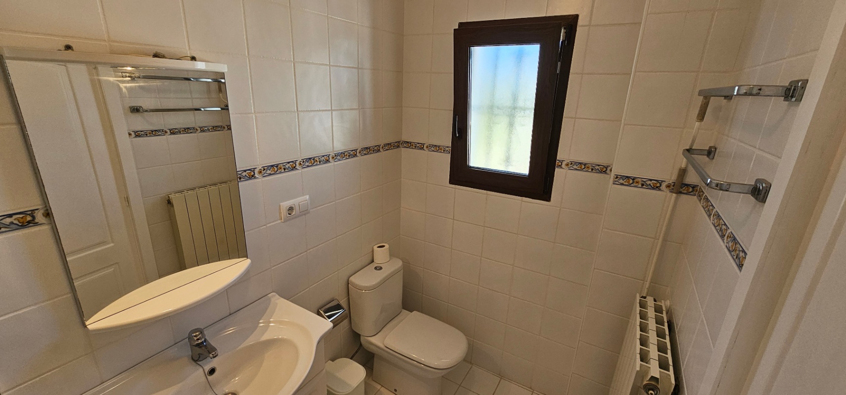 Bathroom featuring white wall tiles with a decorative border, a ceramic toilet, washbasin with mirror, and a dark-framed window for natural light.