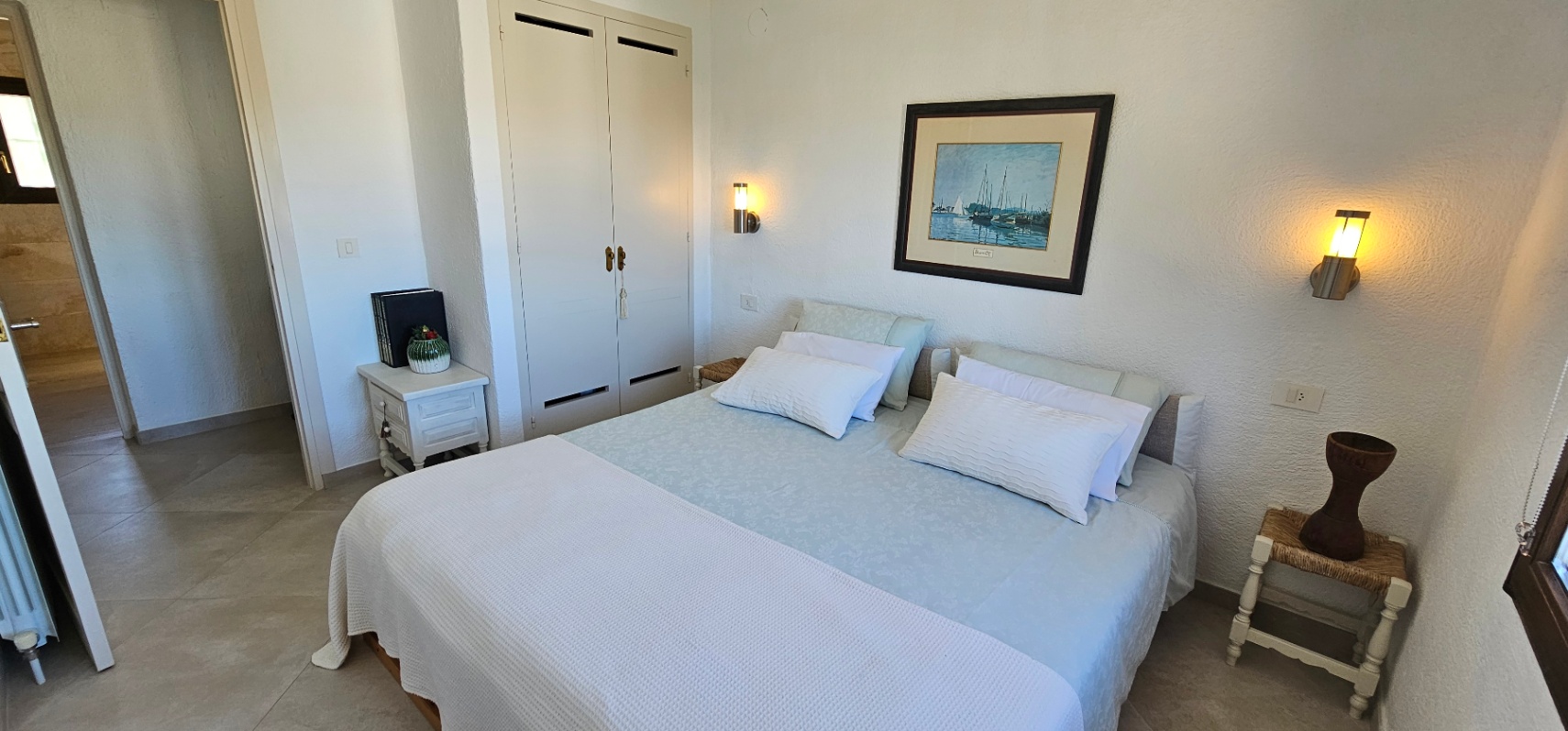 Bedroom featuring a double bed, built-in white wardrobes, and warm wall-mounted lamps. The room includes tiled flooring and textured walls for a clean finish.