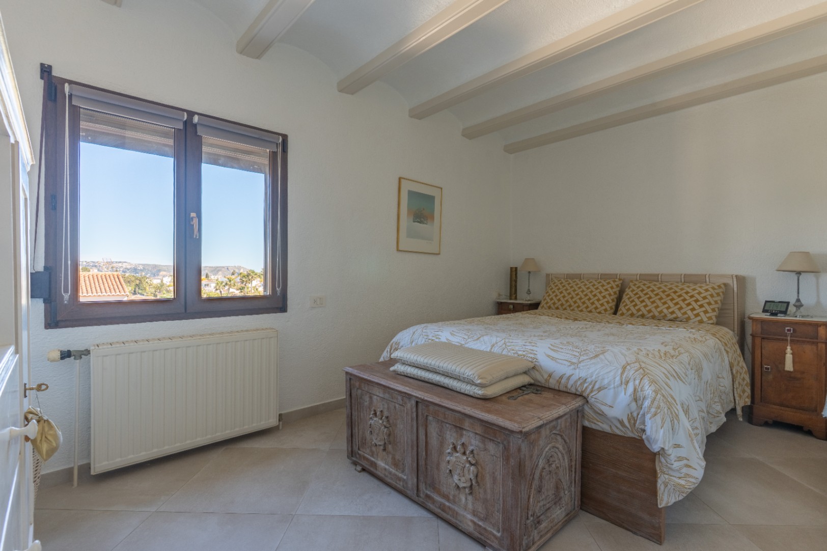Bedroom featuring exposed ceiling beams, ceramic tiles, and a rustic carved chest. It includes central heating and a window overlooking the Jávea landscape.