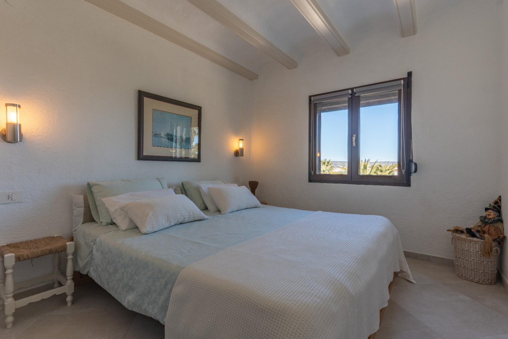 Bright bedroom featuring exposed ceiling beams, modern wall sconces, and a dark wood-framed window overlooking palm trees under a clear blue sky.