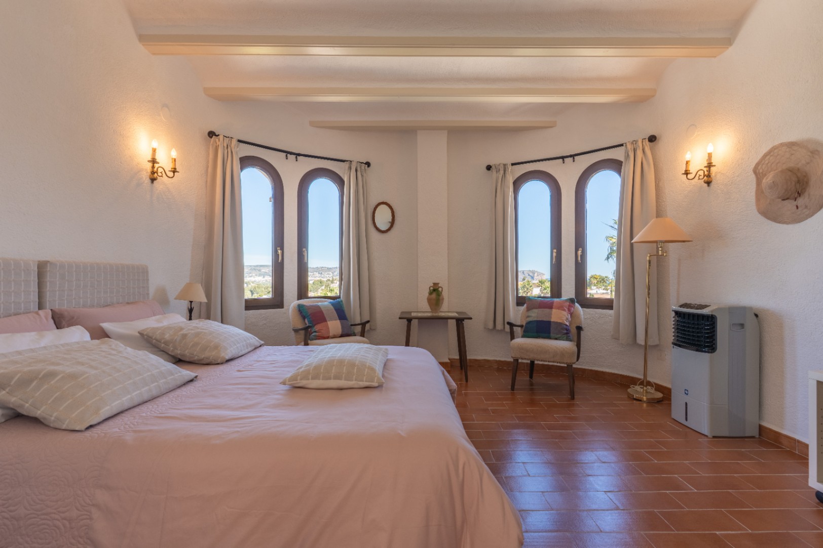 Bright bedroom featuring exposed ceiling beams, terracotta flooring, and arched windows with views of Jávea, complemented by classic candle-style wall sconces.
