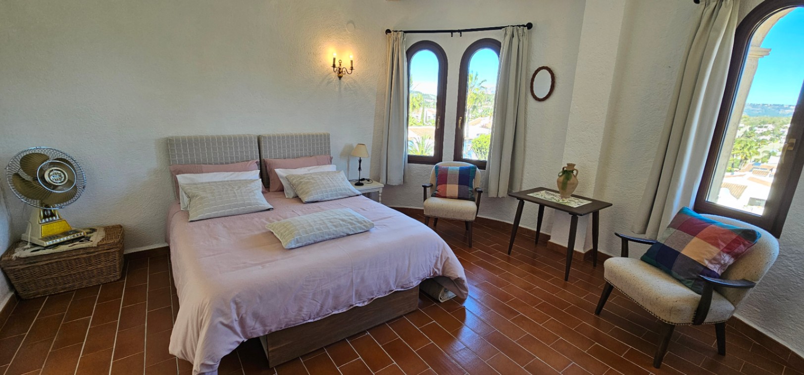 Bright bedroom featuring terracotta tile flooring, traditional arched windows, and a seating area. The space combines rustic charm with natural light and landscape views.