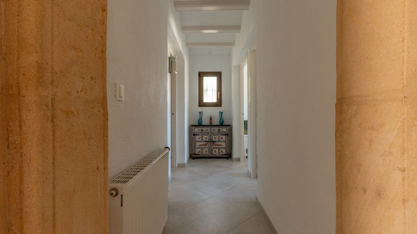 Bright hallway featuring white ceiling beams and traditional Tosca stone pillars. It includes central heating and a decorative chest under a wooden window.