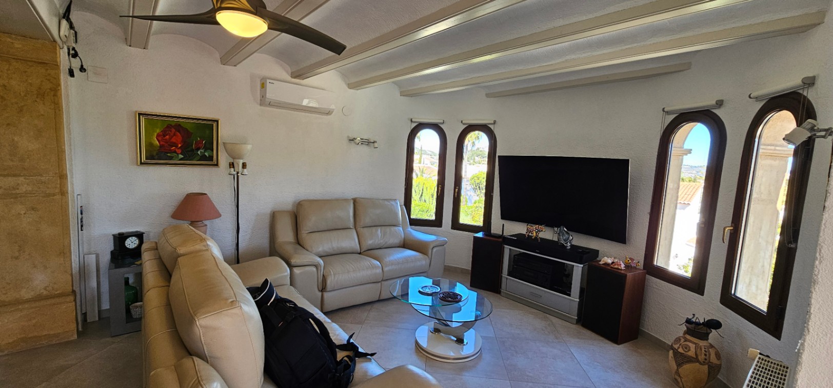 Bright living area featuring beige leather sofas, exposed ceiling beams, air conditioning, and arched windows that provide ample natural light.