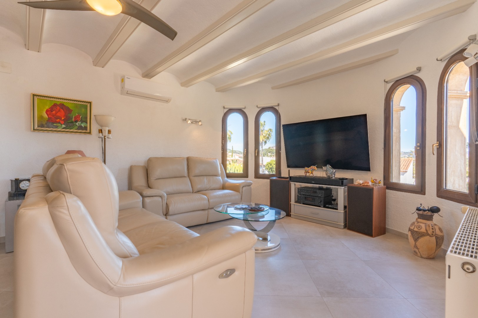 Bright living area featuring exposed ceiling beams, cream leather sofas, and arched windows. Includes air conditioning, a ceiling fan, and a home media setup.