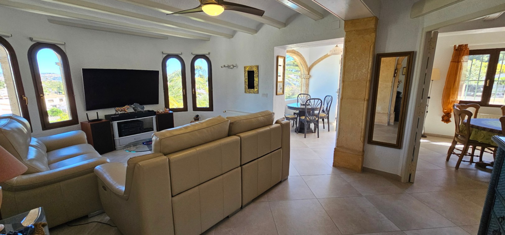 Bright living area featuring white beamed ceilings, beige leather seating, and arched windows. A Tosca stone pillar frames the transition to the dining terrace.