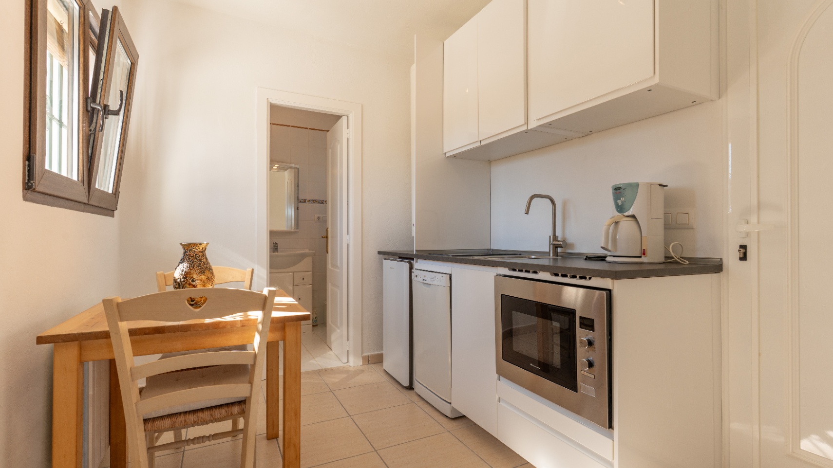 Compact kitchen featuring white cabinetry, integrated appliances, and a wooden dining set, with direct access to an adjacent bathroom.