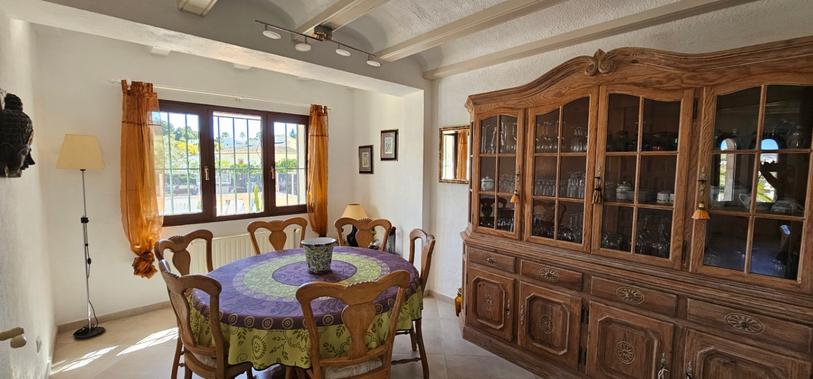 Dining room featuring a round table with carved wooden chairs and a large classic display cabinet. Includes exposed ceiling beams and natural light from the window.