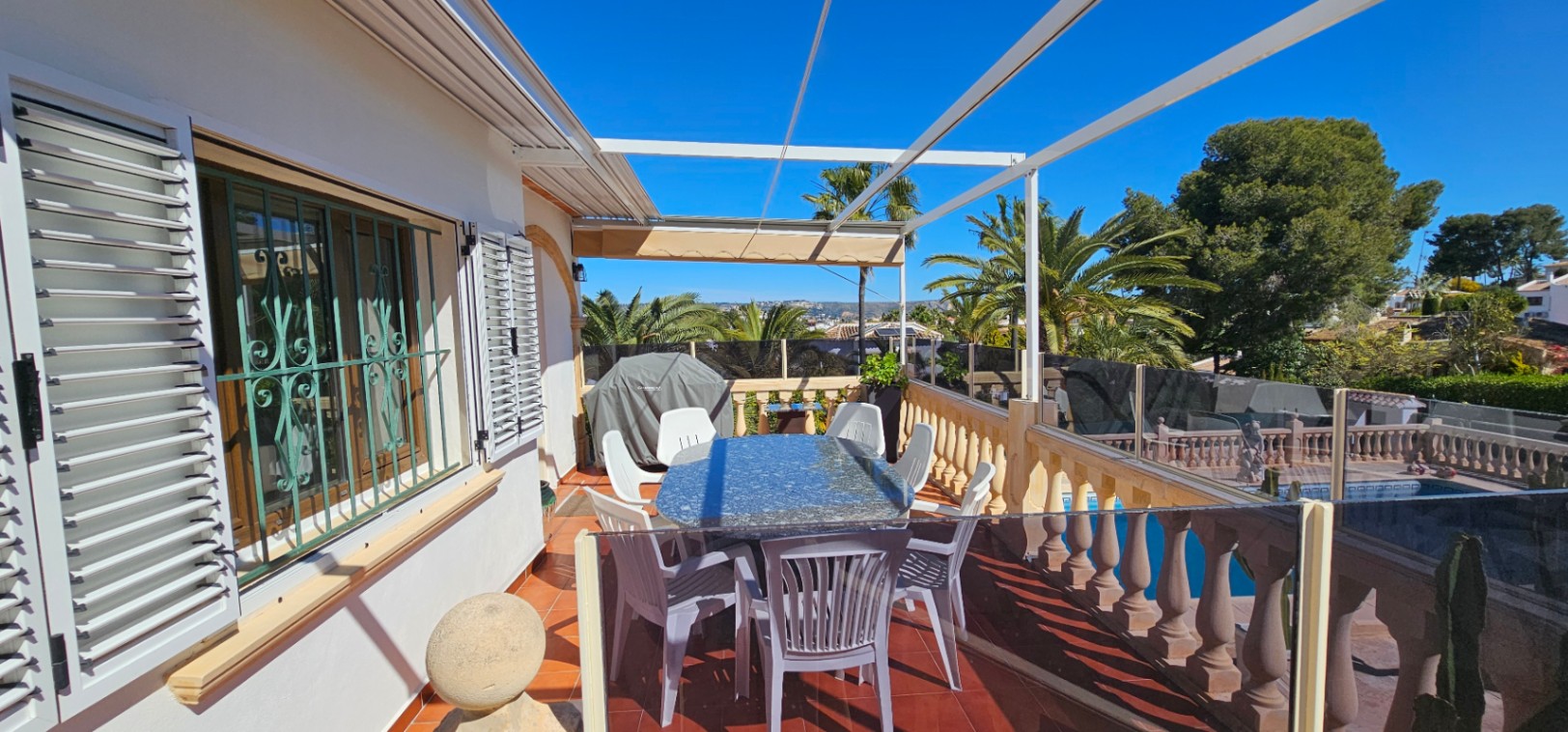 Elevated terrace with terracotta flooring, a granite dining table, and glass balustrades. It overlooks the private pool and lush palm trees under a clear sky.