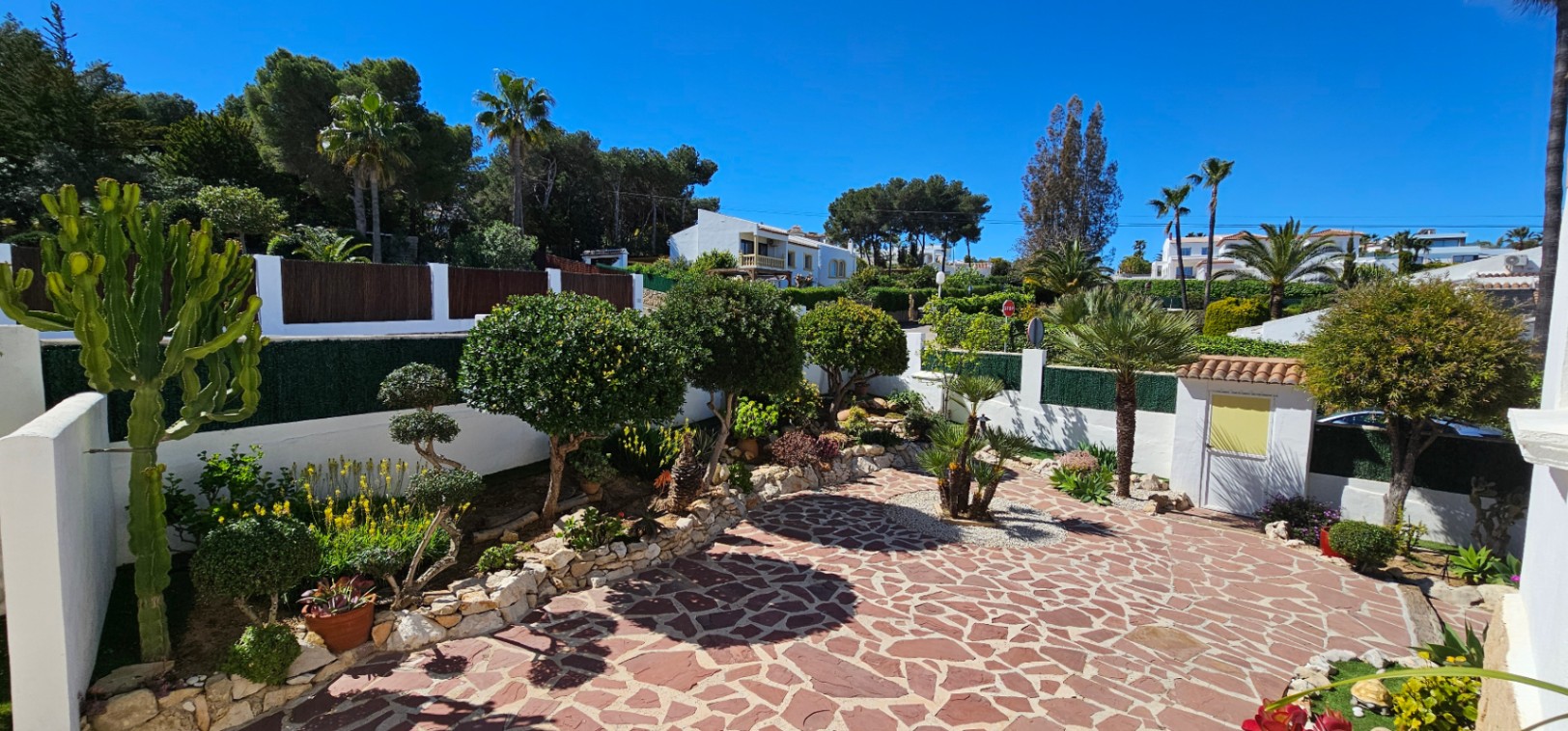 Exterior patio featuring irregular natural stone paving, bordered by raised stone planters with Mediterranean flora, cacti, and palms under clear skies.