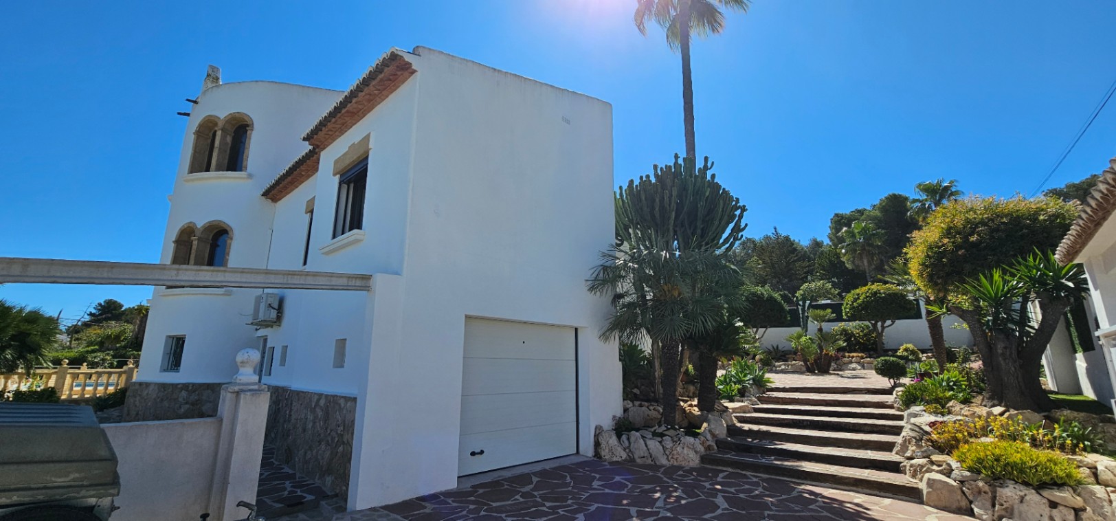 Exterior view of the white villa featuring a stone base, integrated garage, and stone steps leading through a Mediterranean garden with palm trees.