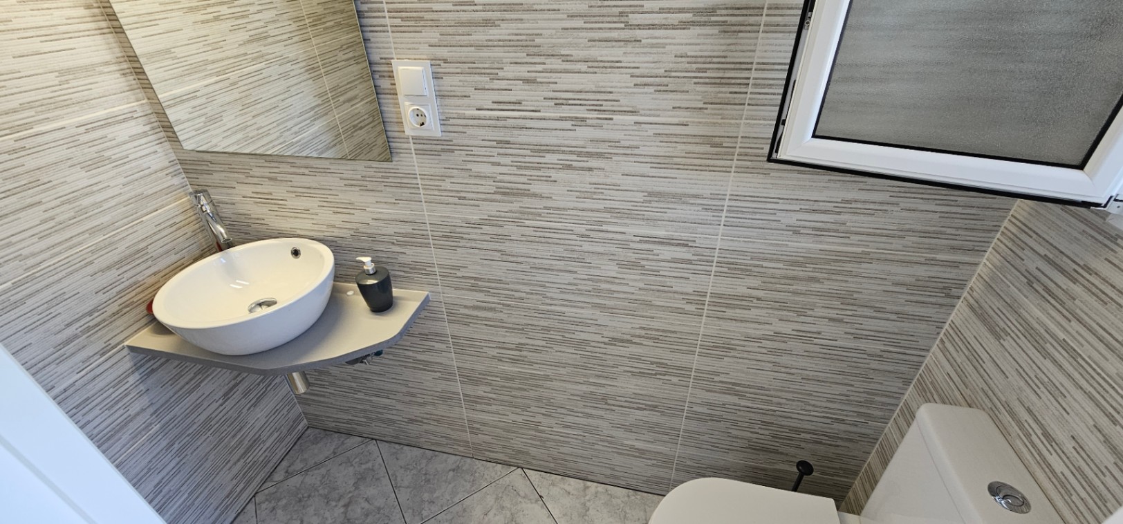 Functional bathroom featuring a circular vessel sink on a grey floating shelf, complemented by textured linear wall tiles and diagonal floor tiling.