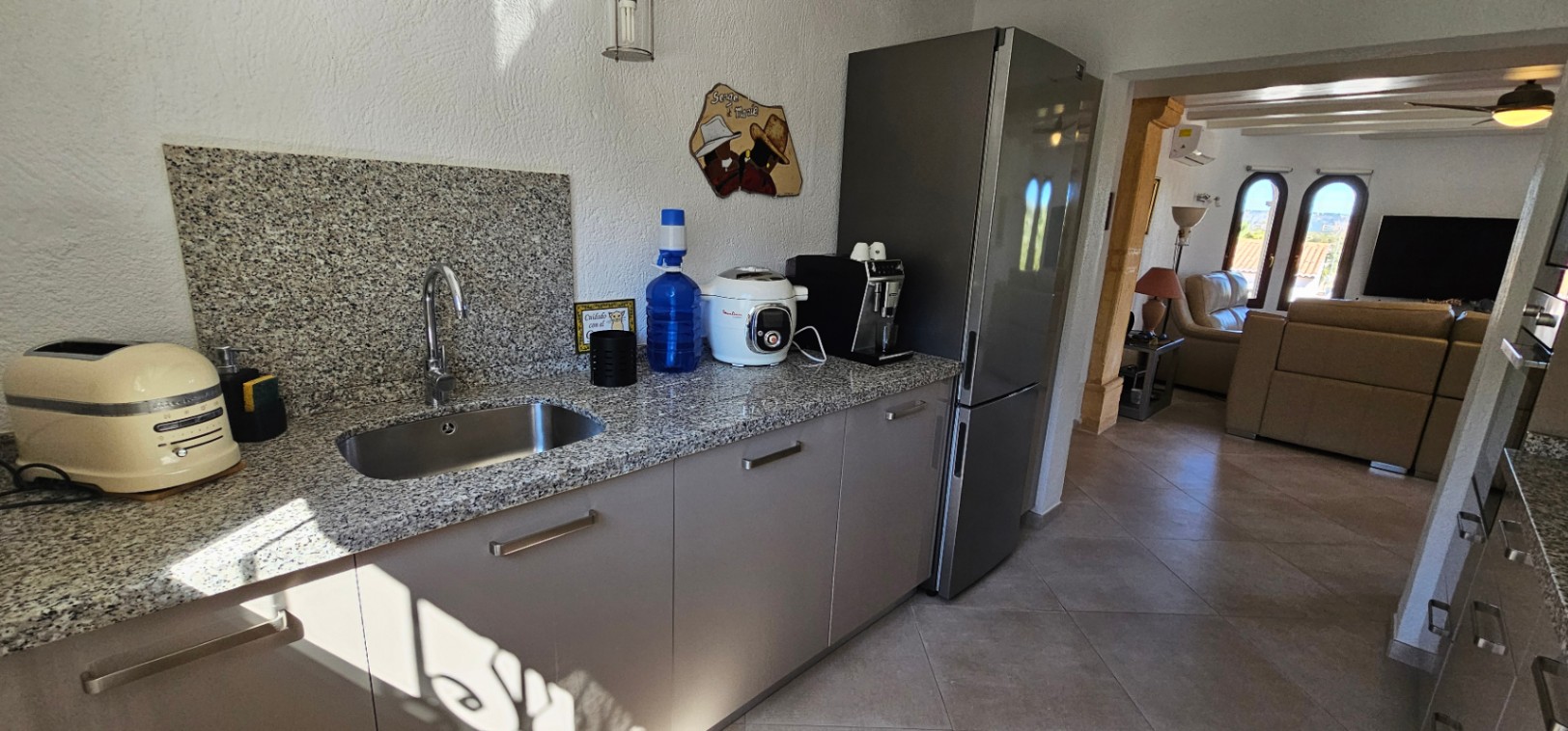Functional kitchen featuring granite countertops and stainless steel sink, opening into a bright living area with leather seating and arched windows.