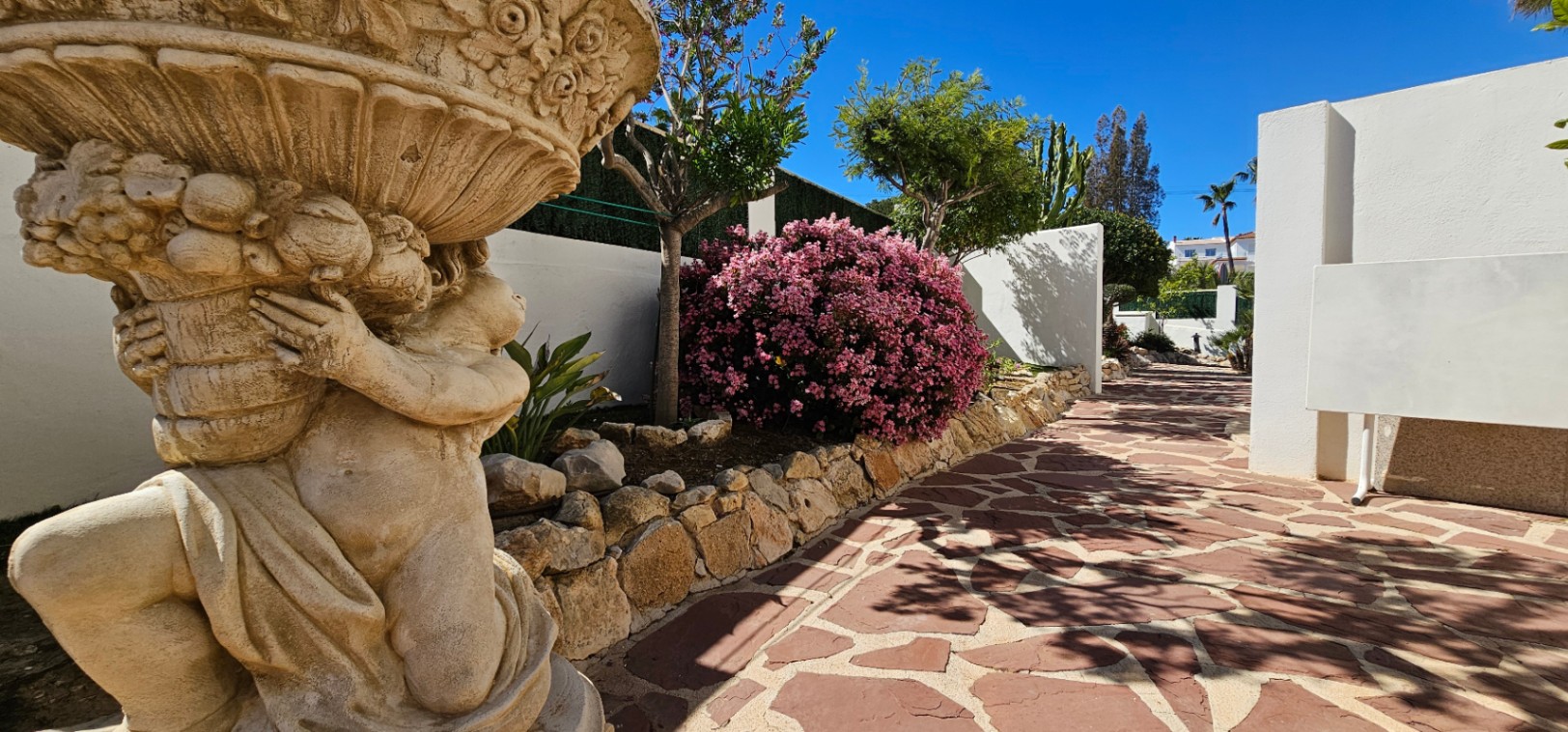 Irregular stone pathway featuring a decorative cherub statue and vibrant Mediterranean flora, including pink oleanders and white perimeter walls.