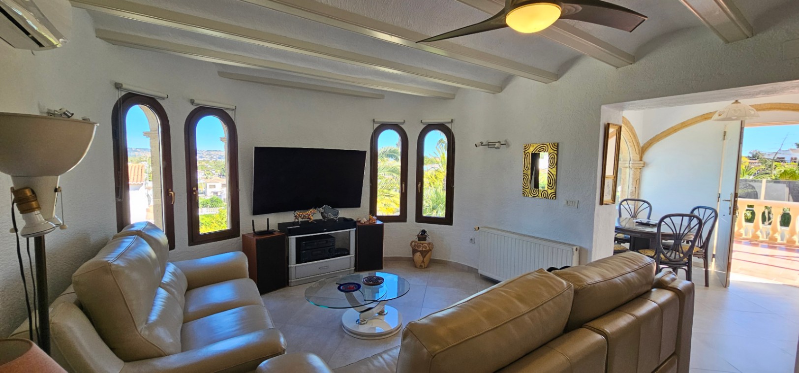 Living area featuring white beamed ceilings, leather sofas, and arched windows. The space connects via a stone archway to the dining room and outdoor terrace.