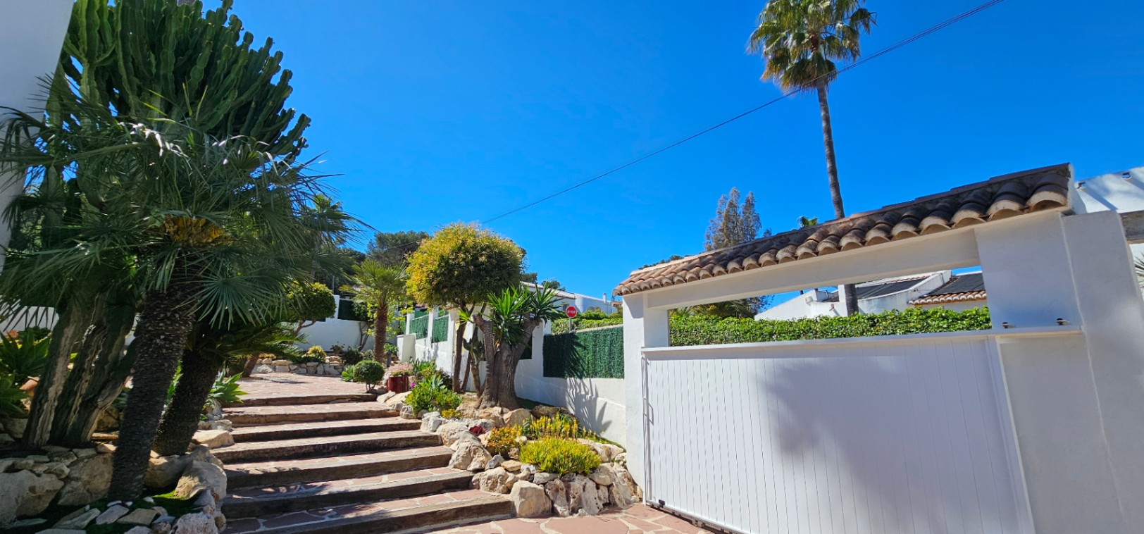 Main entrance featuring a white sliding gate and natural stone steps flanked by mature Mediterranean landscaping, including cacti and palm trees.