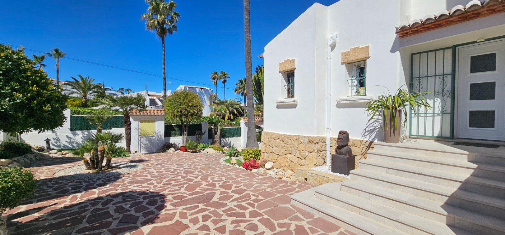 Main entrance featuring wide stone steps, a white facade with natural stone accents, and a paved courtyard surrounded by Mediterranean landscaping and palms.