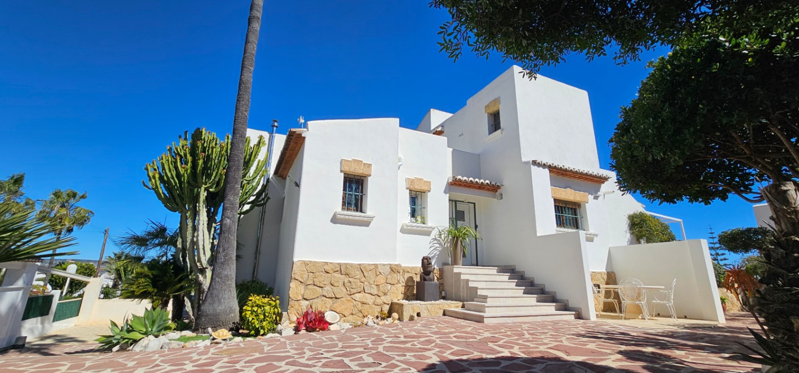 Mediterranean facade featuring white walls, natural stone base, and a stepped entrance. The exterior offers a paved patio surrounded by native cacti and palm trees.