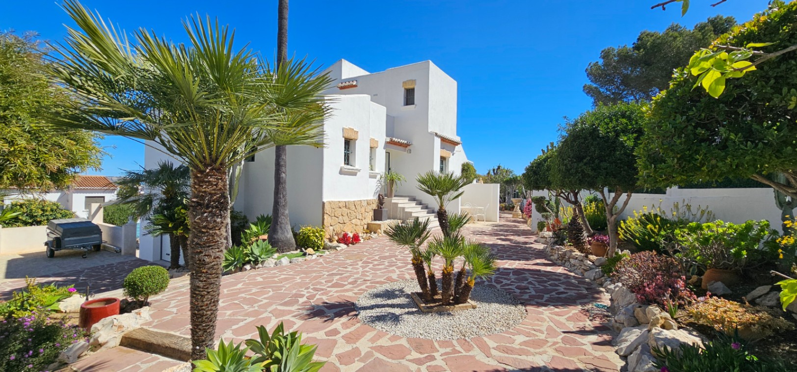 Mediterranean villa exterior featuring a white facade with natural stone accents, landscaped gardens with palms, and a decorative stone-paved driveway.