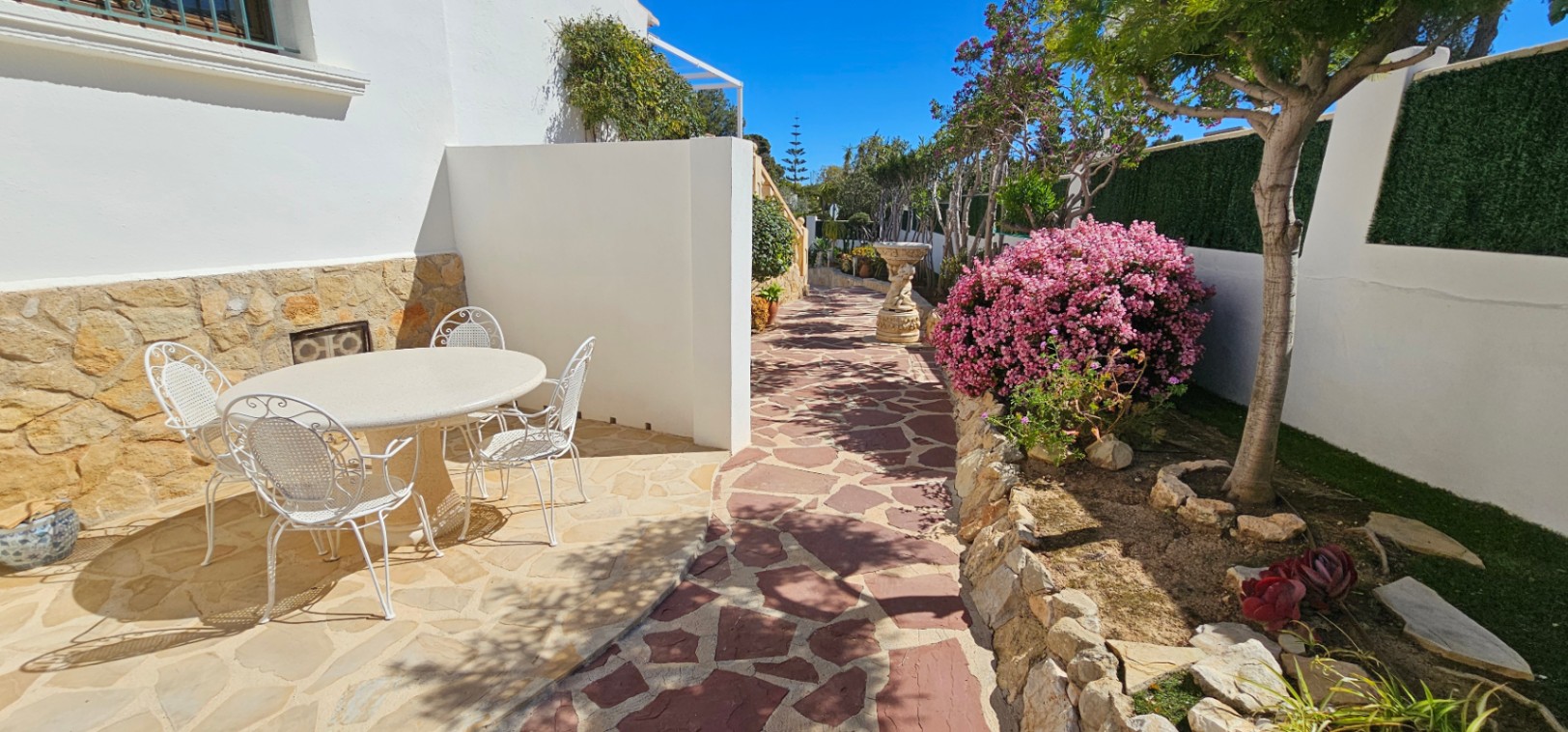 Outdoor patio featuring irregular stone paving, a white wrought-iron dining set, and landscaped gardens with vibrant flowering shrubs and stone borders.